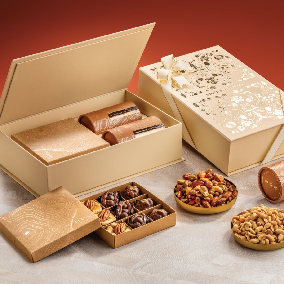 Cream Stories of Bruijn Grand Box with gold floral design, containing two golden cylindrical nut containers, a craft box with assorted confections, and bowls of nuts and chocolates.