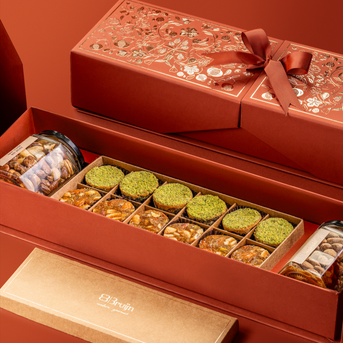 Red Stories of Bruijn Royal Box with gold floral design and ribbon, containing jars of smoked almonds and salted caramel nut mix, alongside a craft box with fig, apricot & nut bites, and pistachio-coated Persian Ranginak.