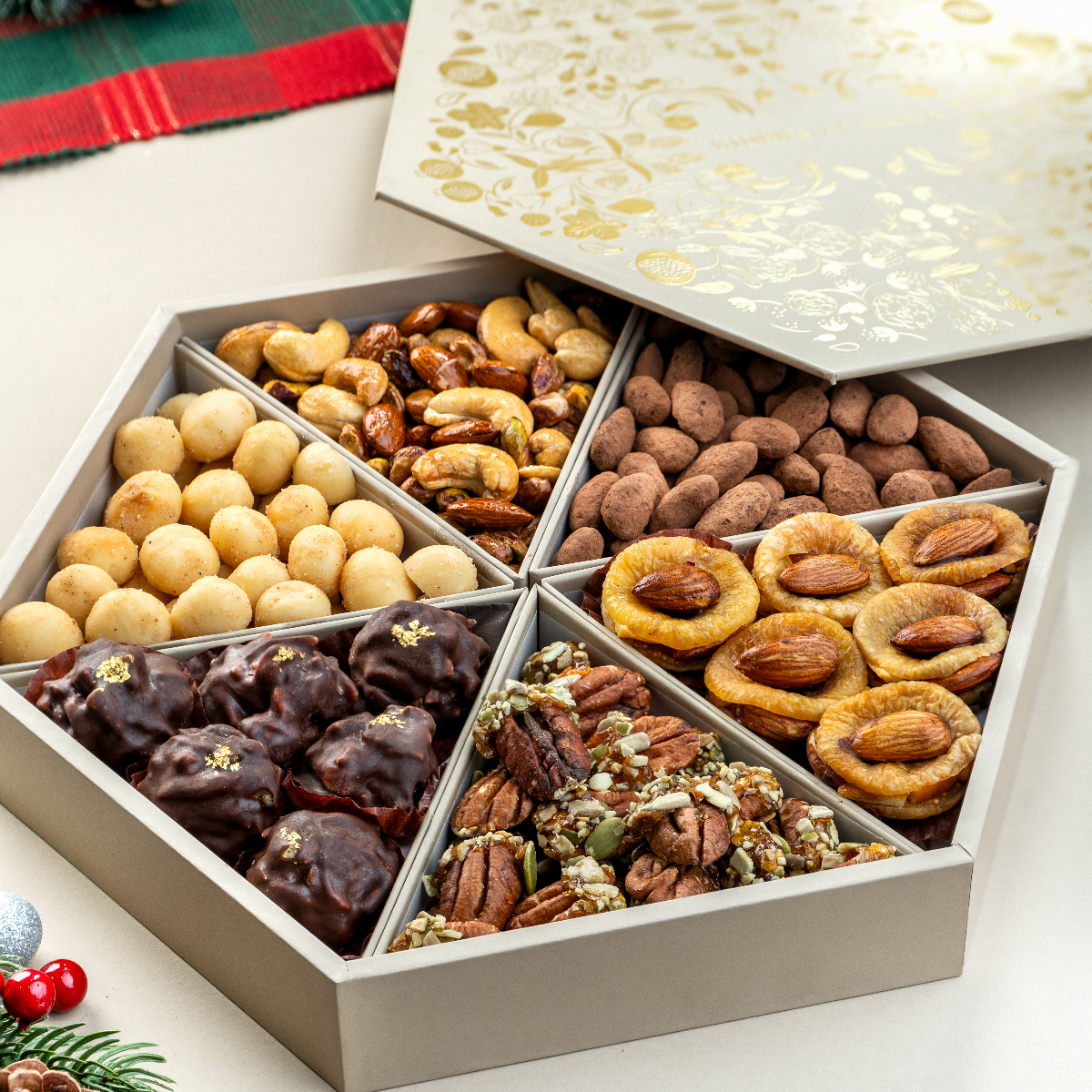 An open medium-sized gift tray containing assorted chocolate and non-chocolate covered nuts and fruits, presented on a festive background with Christmas decorations.