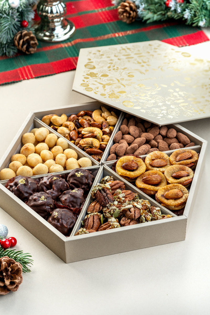 An open medium-sized gift tray containing assorted chocolate and non-chocolate covered nuts and fruits, presented on a festive background with Christmas decorations.