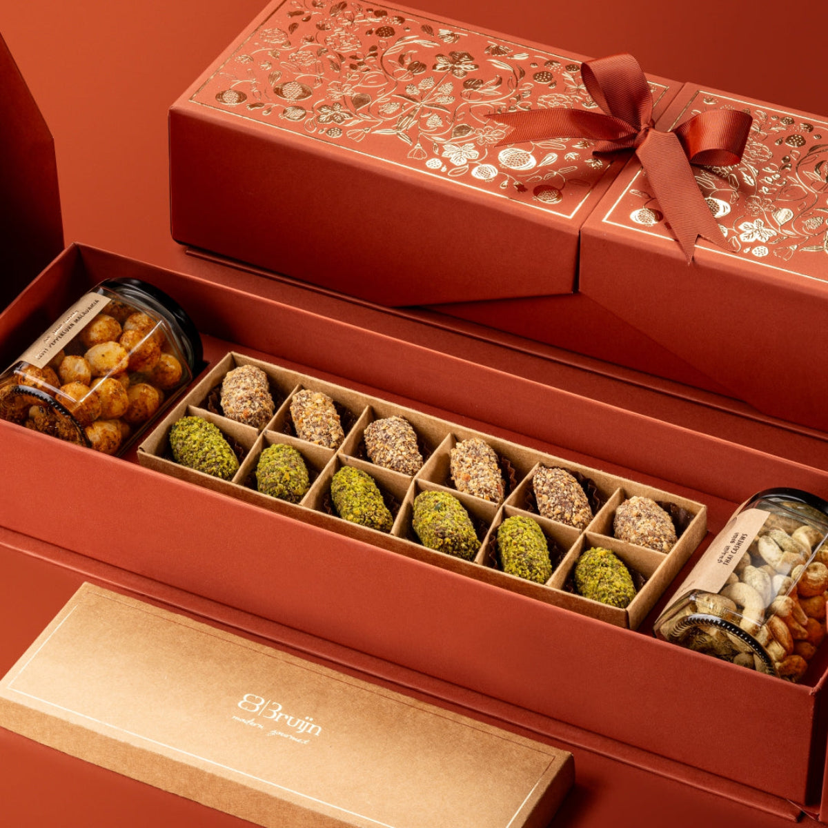 Red Stories of Bruijn Royal Box with gold floral design and ribbon, containing jars of zesty peppercorn macadamias and Thai cashews, alongside a craft box of assorted pistachio, nut, and spice-encrusted date truffles.