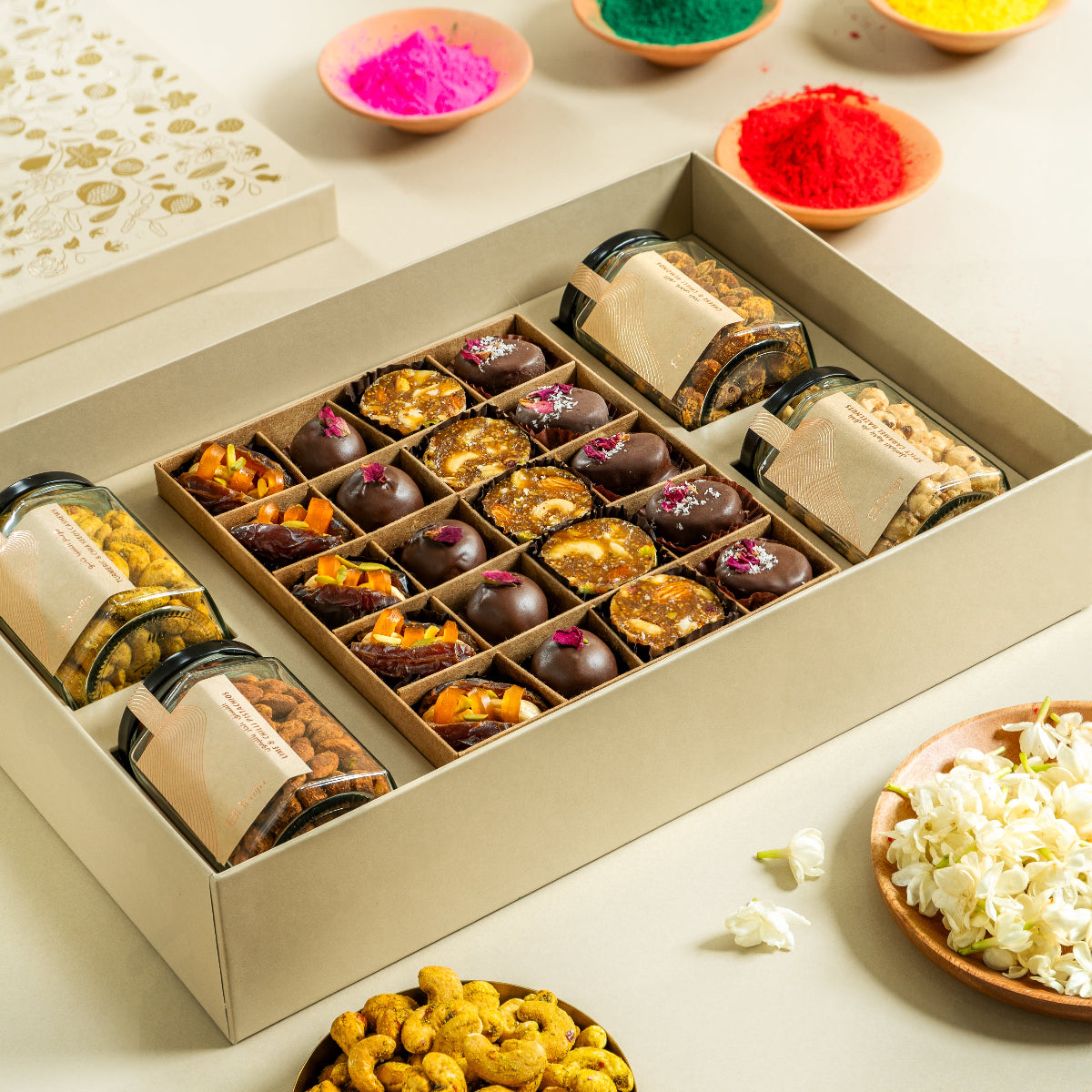 A box of assorted gourmet snacks including nuts, chocolates, and stuffed dates, presented in a festive setting with colorful powders in the background.