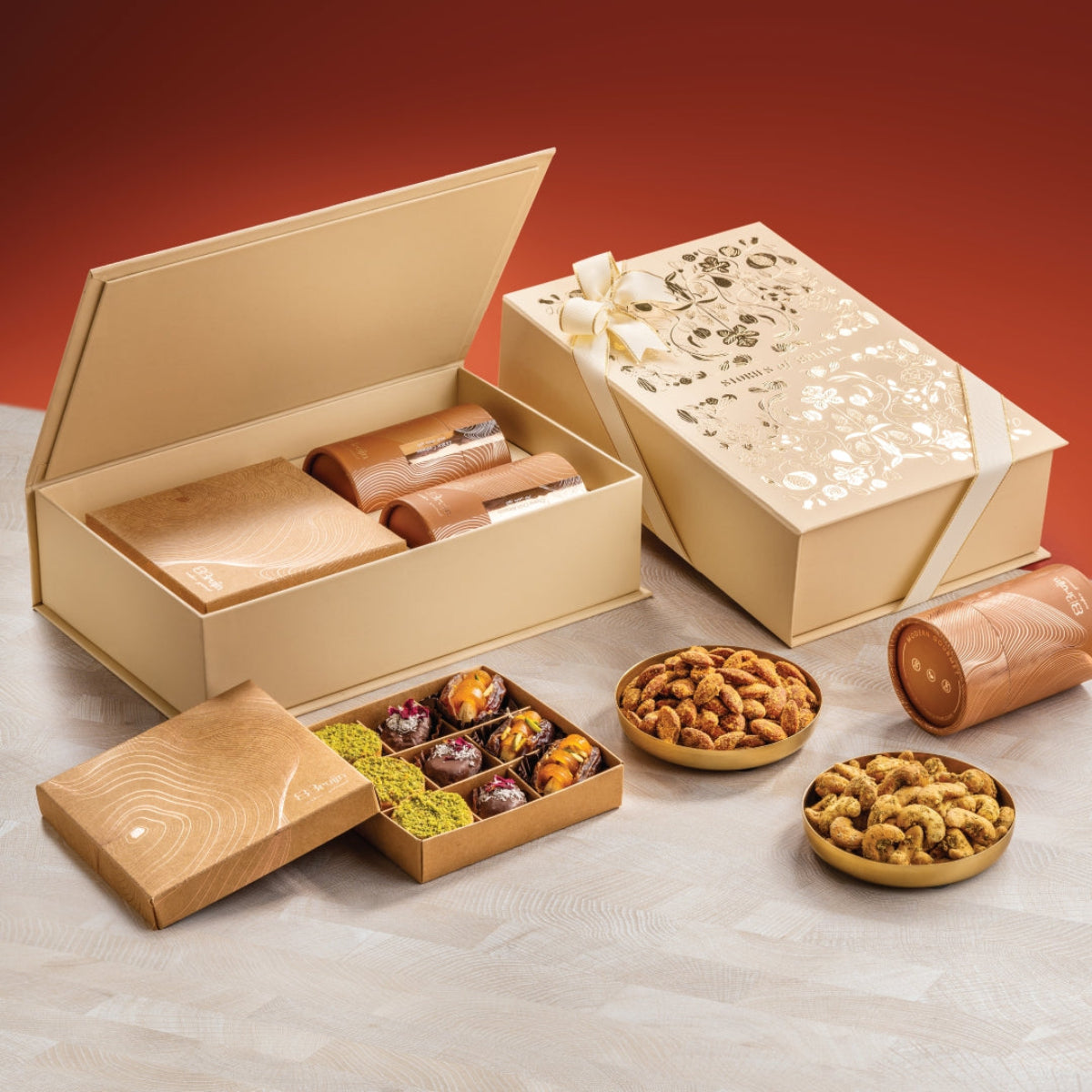 Festive Grand Box with gold floral design and white ribbon. Two golden cylindrical nut containers, a craft box with assorted sweets, and bowls of nuts.