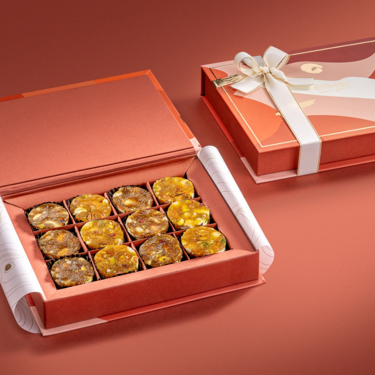 Open Dual Box with patterned lid and ribbon, containing neatly arranged fig, apricot, and date nut bites in golden brown shades.