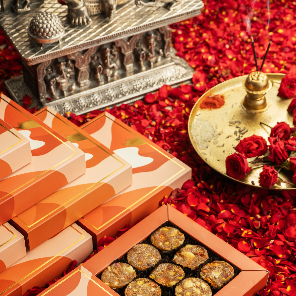 A box of Fig & Nut Bites in Petite Box, displayed alongside a silver metal box and a small lamp with incense on a red background, with scattered rose petals around.