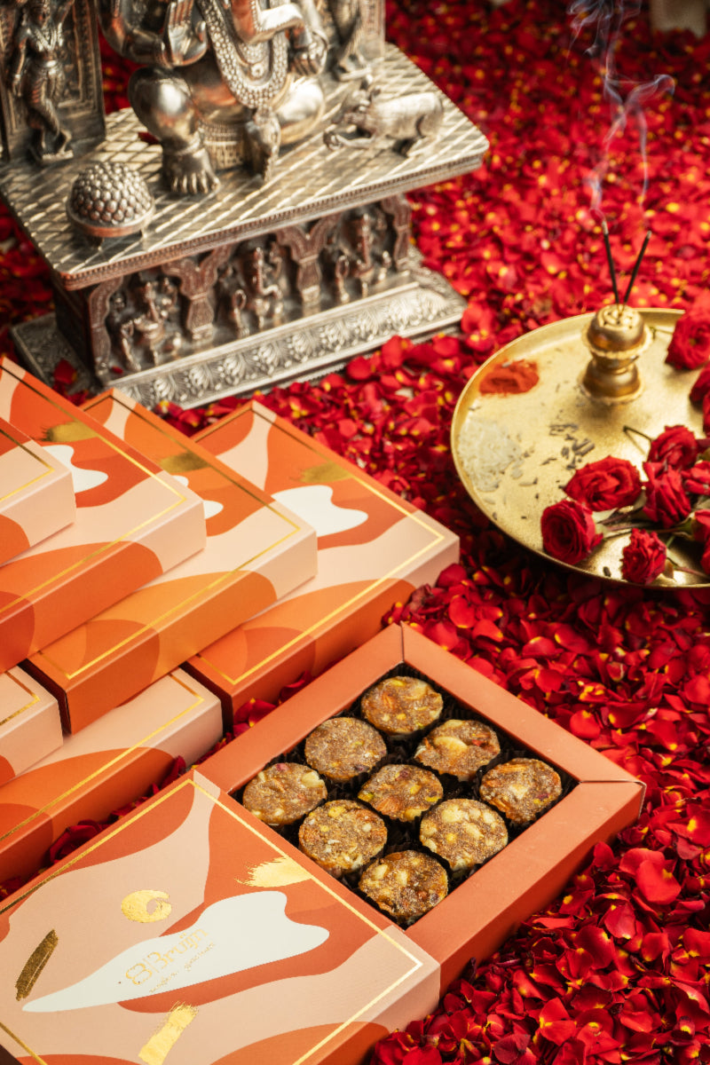 A box of Fig & Nut Bites in Petite Box, displayed alongside a silver metal box and a small lamp with incense on a red background, with scattered rose petals around.