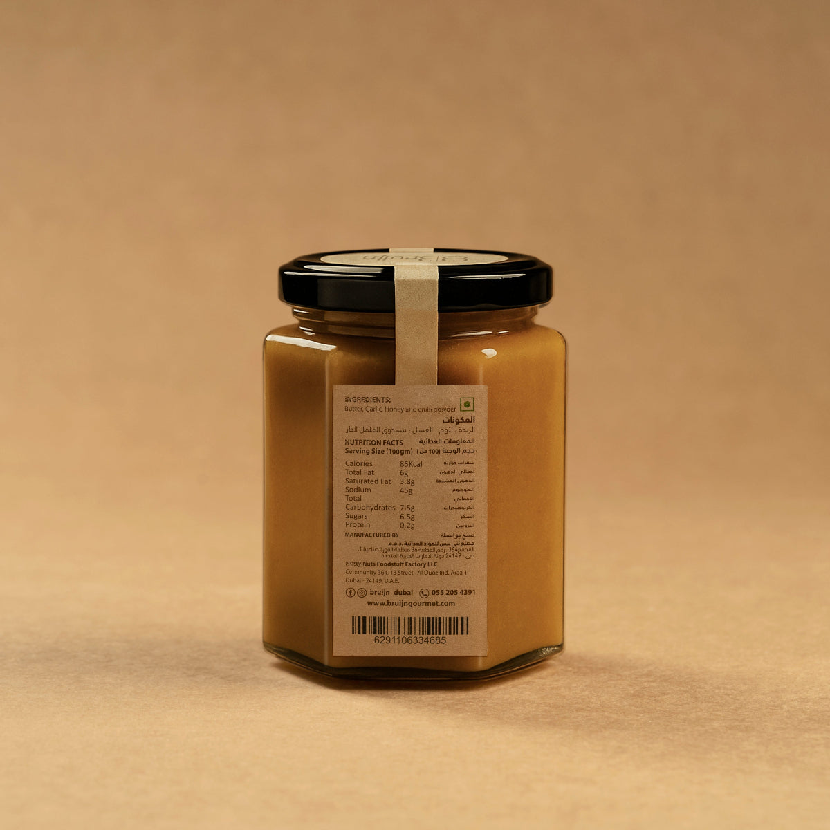 Glass jar of Bruijn Hot Honey Butter with black lid and kraft label, filled with a golden-red blend of garlic, red chillis, and raw honey.
