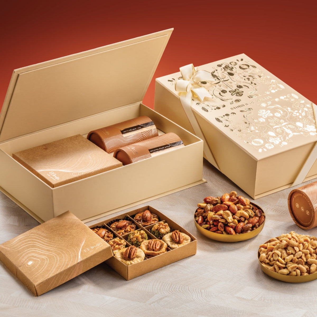 Cream Stories of Bruijn Grand Box with gold floral design, containing two golden cylindrical nut containers, a craft box with assorted fig and nut confections, and bowls of nuts.