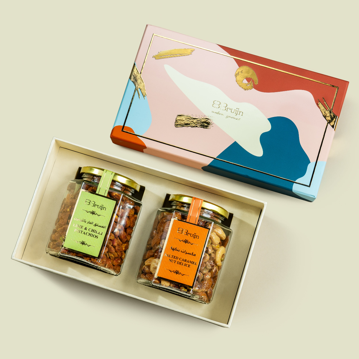 Open Modern Abundance Box with colorful abstract lid, showcasing jars of Salted Caramel Nut Délice and Lime & Chilli Pistachios kernels with gold lids and bright labels.