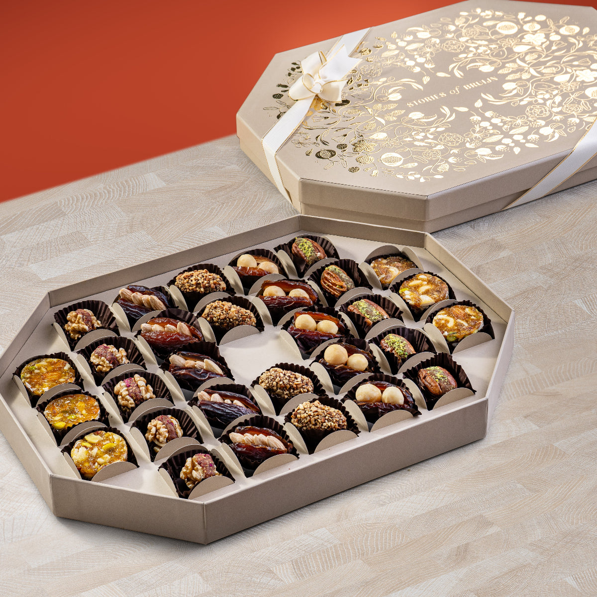 Elegant cream Ruby Box with gold floral design and ribbon, filled with an assortment of fig & nut bites, almond and macadamia stuffed dates, tamriyas, pecan wholes, and apricot nut bites.