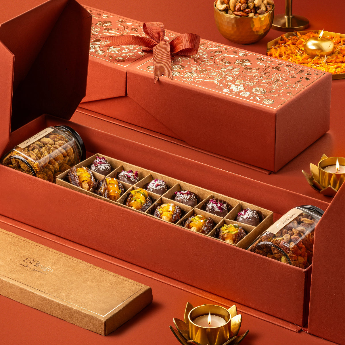 Red Stories of Bruijn Royal Box with gold floral design and ribbon, containing jars of lime & chilli pistachios and cheese & chilli almonds, alongside a craft box with chocolate-coated paan bites and mango cashew stuffed dates.