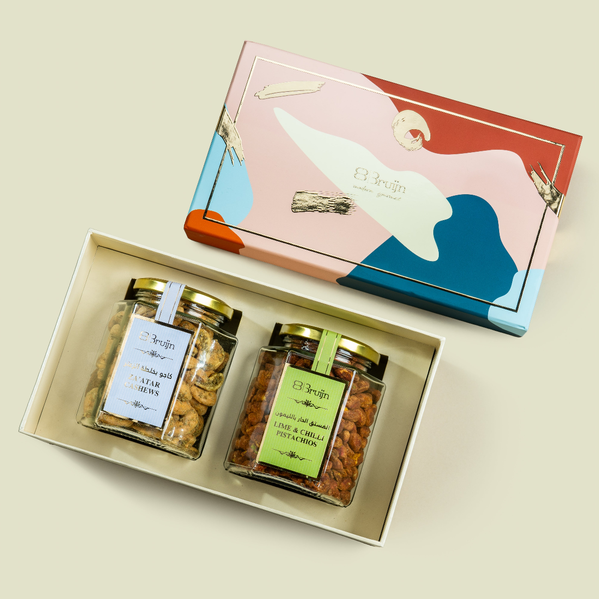 Open Modern Abundance Box with colorful abstract lid, showcasing two jars of Za’atar Cashews and Lime & Chilli Pistachios kernels with gold lids and labels.