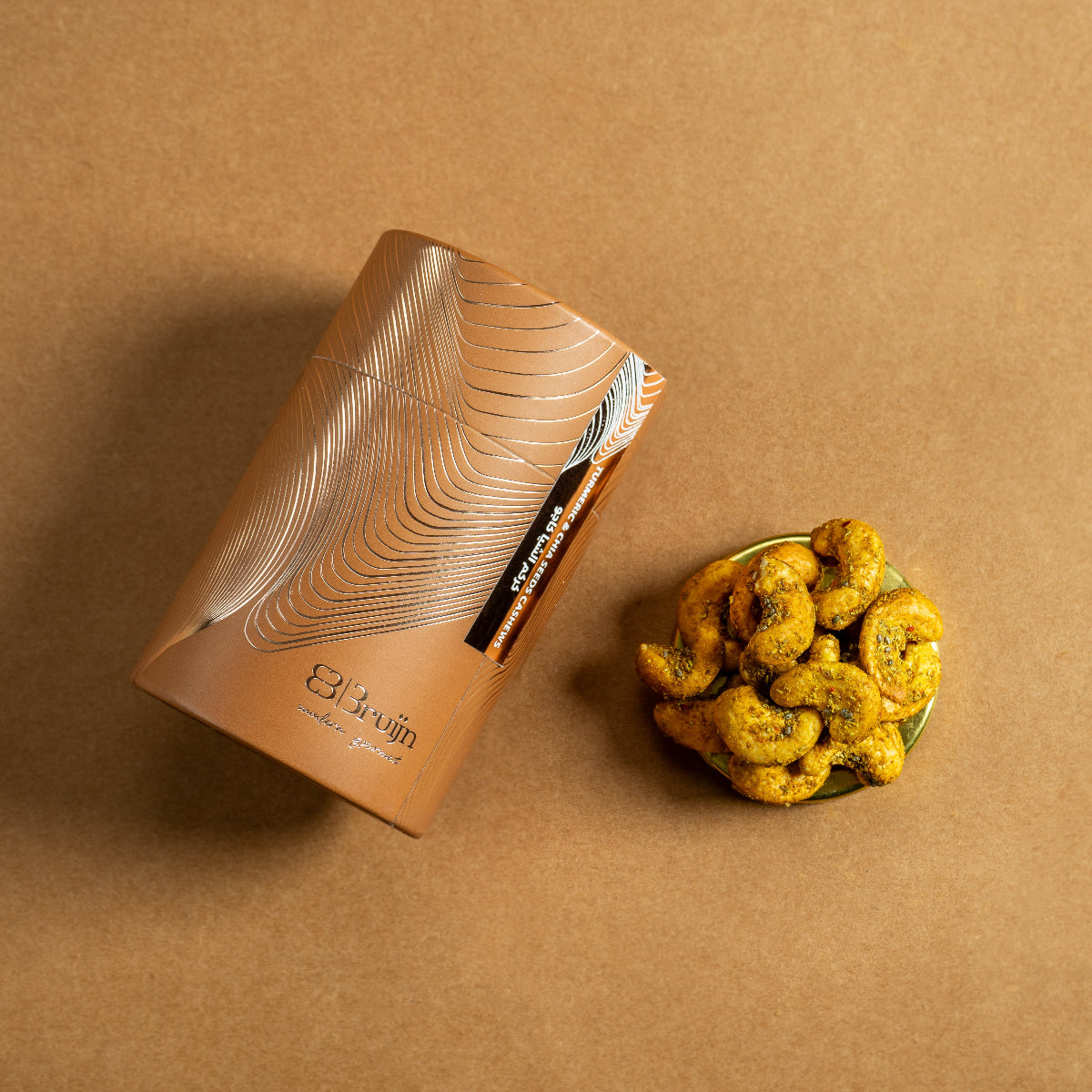 Brown cylindrical container of Bruijn Turmeric & Chia Seed Cashews with minimalist design, containing golden turmeric-coated cashews speckled with chia seeds.
