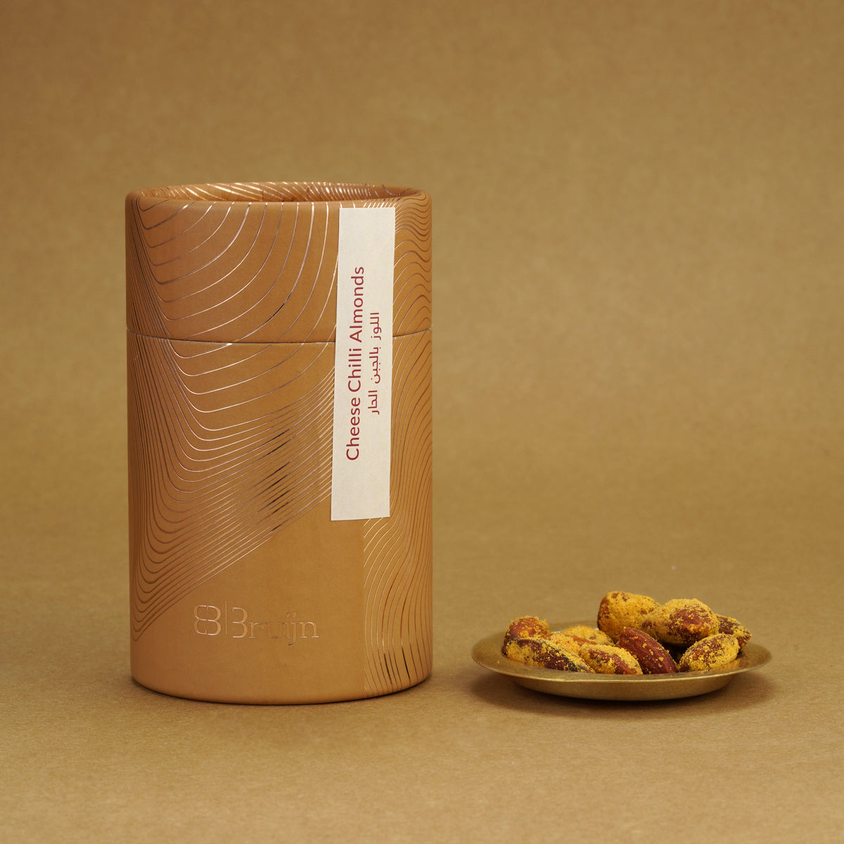 Brown cylindrical container of Bruijn Cheese & Chilli Almonds with minimalist design, placed beside a plate of golden almonds coated in cheese and chilli seasoning.