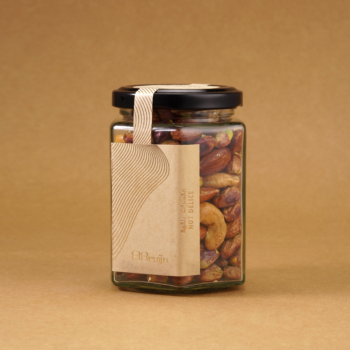 Glass jar of Bruijn Salted Caramel Nut Délice with black lid and elegant kraft label, filled with cashews, almonds, and pistachios coated in golden caramel.
