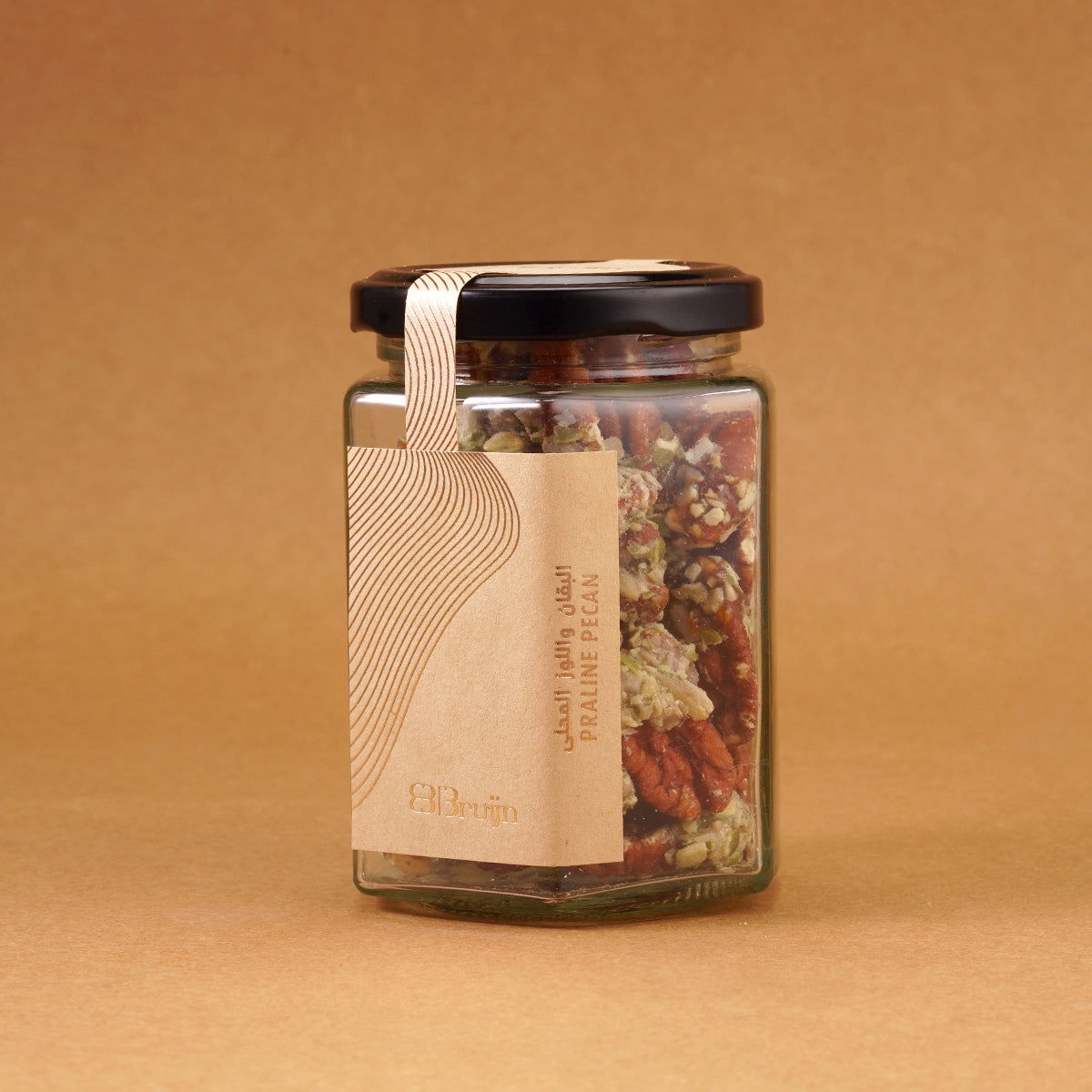 Glass jar of Bruijn Praline Pecans with black lid and kraft label, filled with caramel-coated pecans encrusted with roasted seeds for a sweet and crunchy treat.
