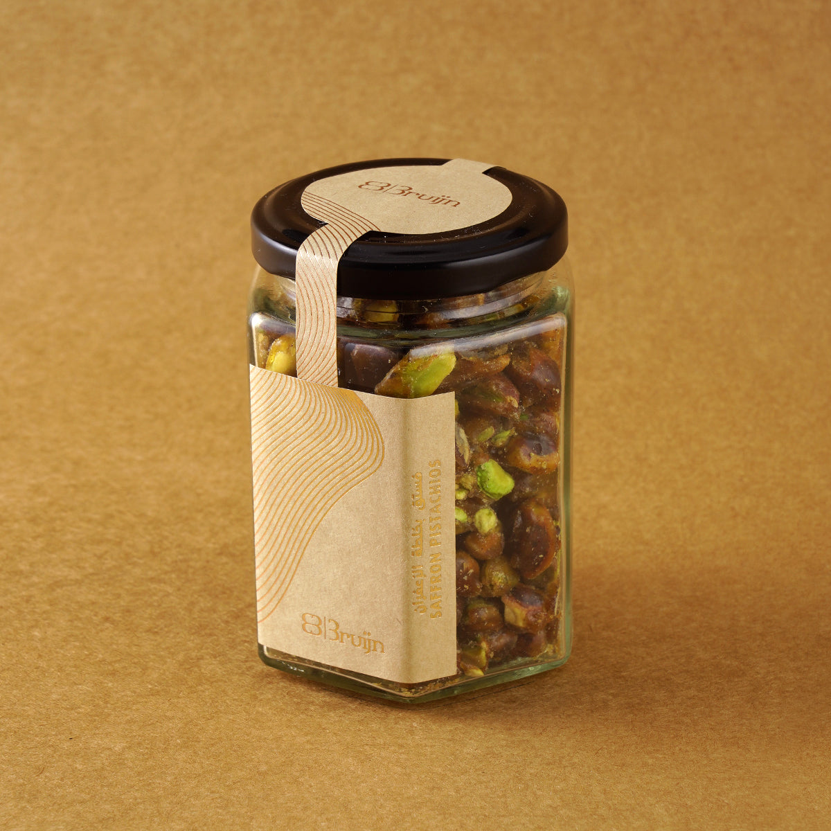 Glass jar of Bruijn Saffron Caramel Pistachios with black lid and kraft label, filled with golden caramel-coated pistachio kernels infused with saffron strands.