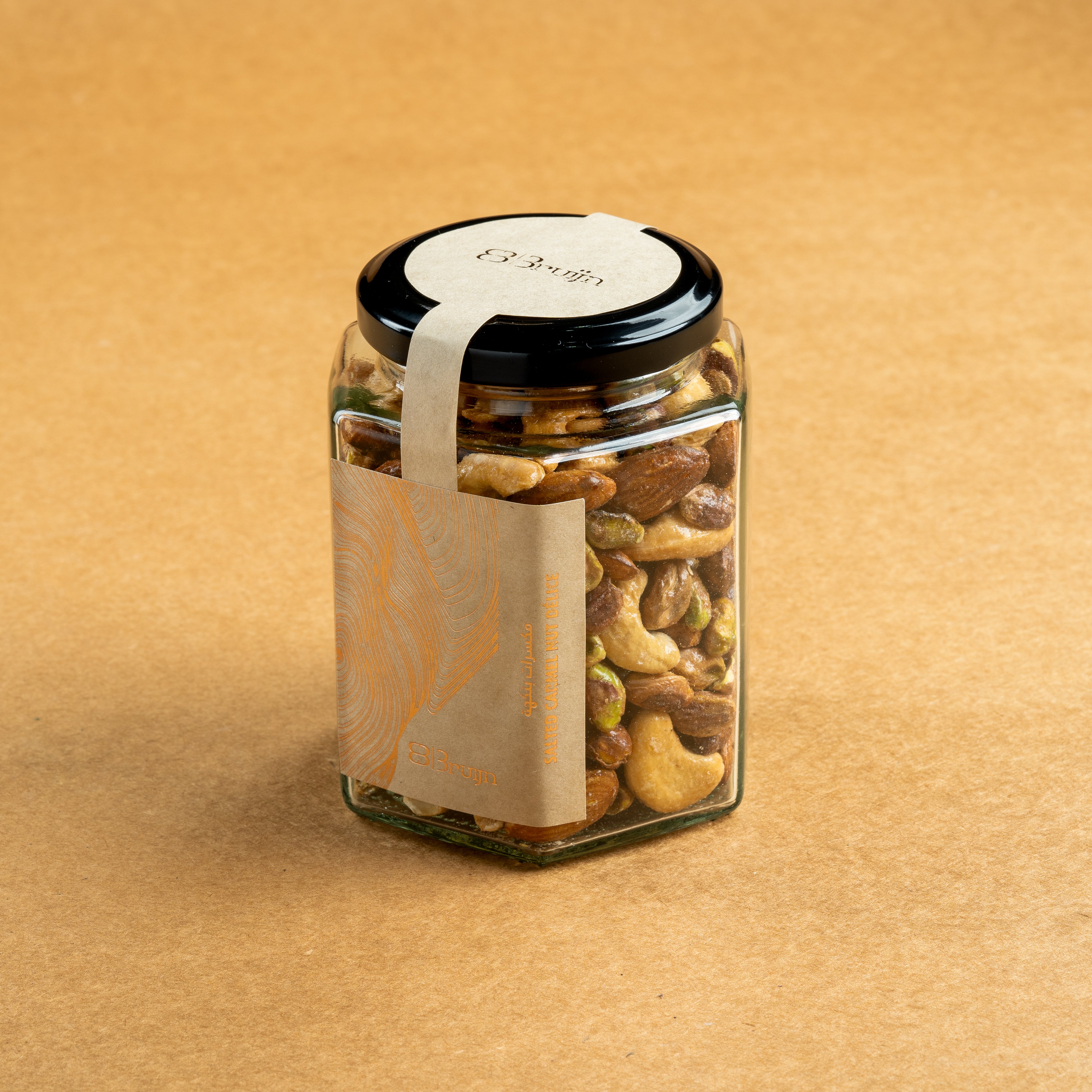 Glass jar of Bruijn Salted Caramel Nut Délice with black lid and elegant kraft label, filled with cashews, almonds, and pistachios coated in golden caramel.