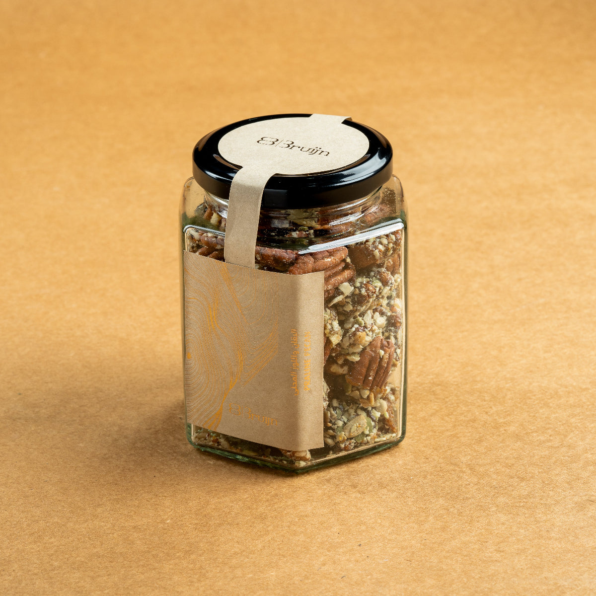 Glass jar of Bruijn Praline Pecans with black lid and kraft label, filled with caramel-coated pecans encrusted with roasted seeds for a sweet and crunchy treat.