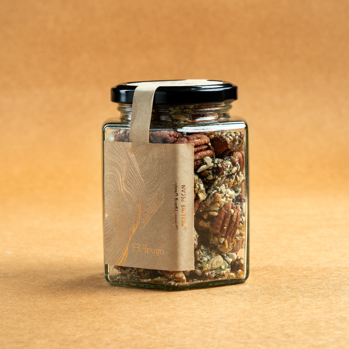 Glass jar of Bruijn Praline Pecans with black lid and kraft label, filled with caramel-coated pecans encrusted with roasted seeds for a sweet and crunchy treat.