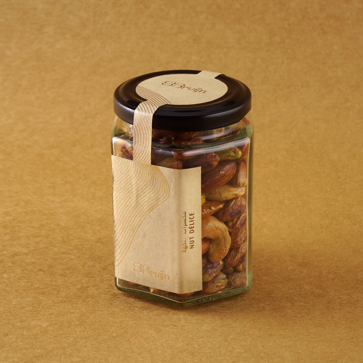Glass jar of Bruijn Salted Caramel Nut Délice with black lid and elegant kraft label, filled with cashews, almonds, and pistachios coated in golden caramel.